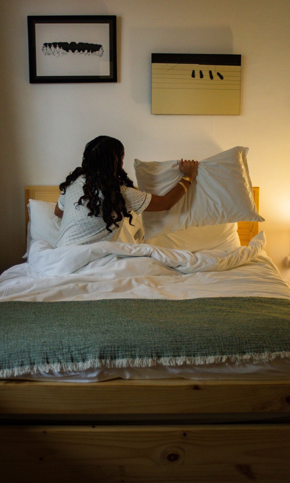 Guest fluffing pillows on a white bed beneath framed art in warm lamplight